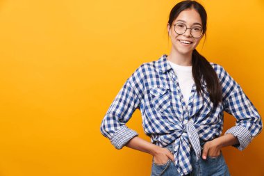 İyimser, mutlu, gözlüklü genç kız sarı duvar arkasında izole bir şekilde poz veriyor..