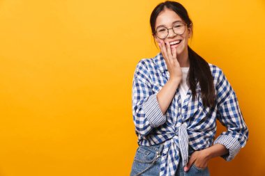 İyimser, mutlu, gözlüklü genç kız sarı duvar arkasında izole bir şekilde poz veriyor..