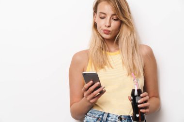 Düşünceli, genç ve güzel bir kadın beyaz duvar arkasına izole edilmiş bir şekilde poz veriyor. Cep telefonuyla soda içiyor..