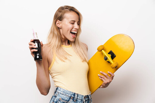 Очаровательная красивая молодая женщина позирует на белом фоне, держа скейтборд, пьющий газировку.
.