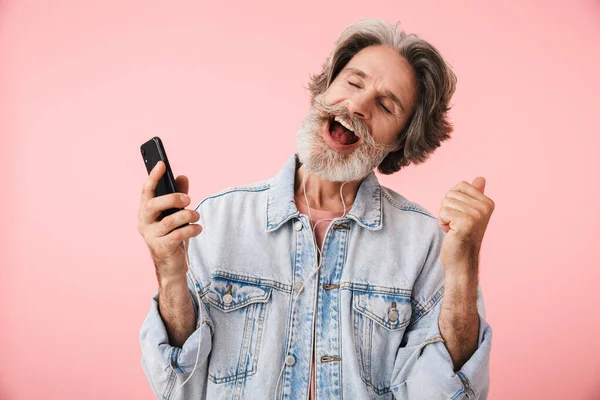 Portrait closeup of happy old man 70s with gray beard singing while listening to music with smartphone and earphones