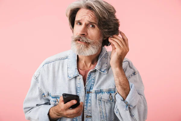 Portret close-up van serieuze oude man jaren 70 met grijze baard houden smartphone tijdens het luisteren naar muziek met oortelefoons — Stockfoto