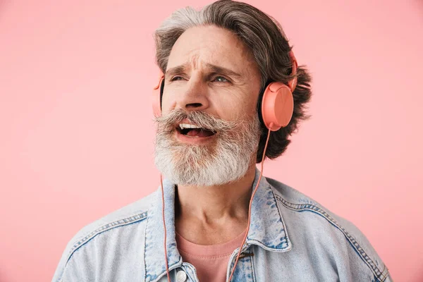 Portrait closeup of cheerful old man 70s with gray beard singing while listening to music with headphones