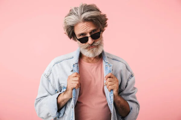 Portret zbliżenie moda stary człowiek 70 z szarą brodą patrząc w aparat fotograficzny i dotykając jego dżinsową kurtkę — Zdjęcie stockowe