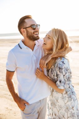 Mutlu bir çiftin güneşli sahilde yürürken gülüp kucaklaştığı bir fotoğraf.