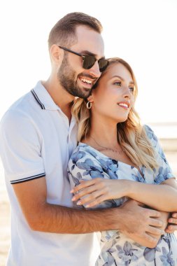 Güneşli sahilde yürürken gülümseyen ve sarılan güzel çiftin fotoğrafı.