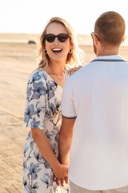 Güneşli sahilde yürürken birlikte gülüp el ele tutuşan mutlu çiftin fotoğrafı.