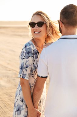 Güneşli sahilde yürürken gülümseyen ve el ele tutuşan mutlu çiftin fotoğrafı.