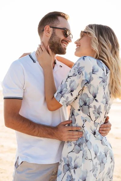 Foto de pareja joven feliz hombre y mujer sonriendo y abrazándose mientras caminan en la playa soleada — Foto de Stock