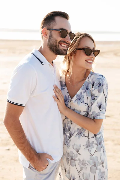 Fotografie usmívající se mladý pár muž a žena dívá stranou a objímání při chůzi na slunné pláži — Stock fotografie