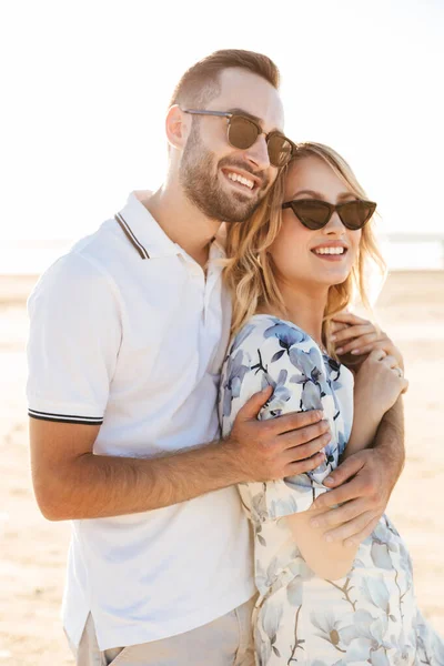 Φωτογραφία του χαμογελαστού καυκάσιου ζευγαριού άντρας και γυναίκα που κοιτούν στην άκρη και αγκαλιάζονται ενώ περπατούν στην ηλιόλουστη παραλία — Φωτογραφία Αρχείου