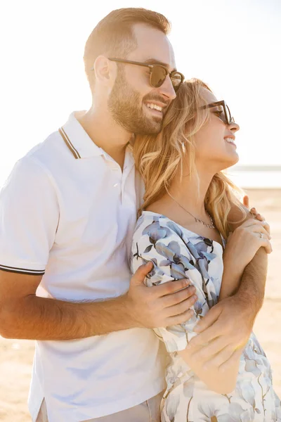 Foto eines lächelnden attraktiven Paares mit Sonnenbrille, das zur Seite schaut und sich beim Spazierengehen am sonnigen Strand umarmt — Stockfoto