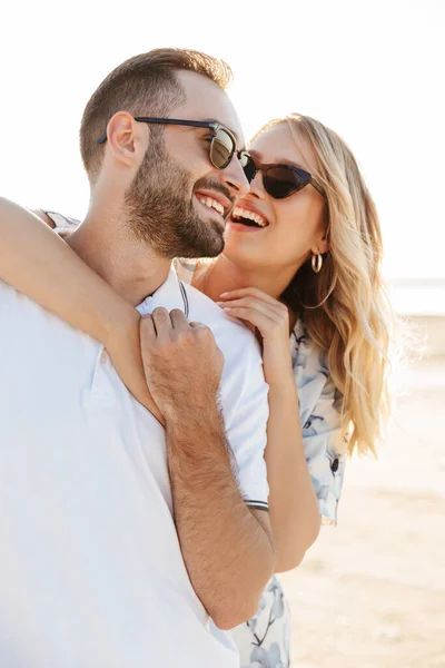 Foto de pareja joven y feliz con gafas de sol riéndose y abrazándose mientras camina en la playa soleada — Foto de Stock