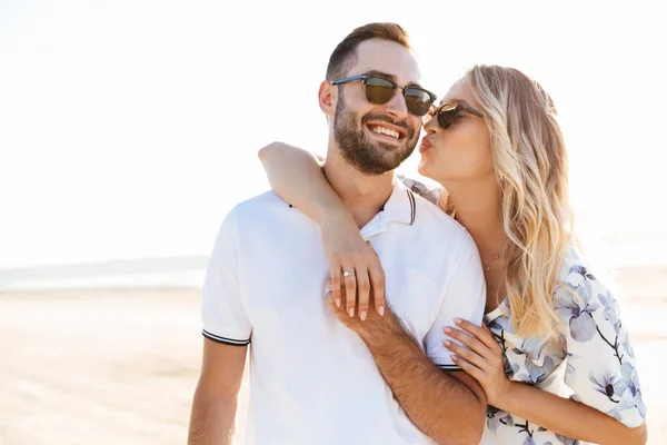 Foto de jovem loira beijando e abraçando homem bonito enquanto caminhava na praia ensolarada — Fotografia de Stock