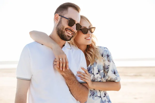 Photo de heureux beau couple portant des lunettes de soleil souriant et étreignant tout en marchant sur la plage ensoleillée — Photo