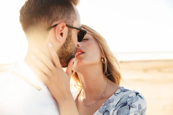 Foto de pareja hermosa romántica que usa ropa de verano besándose y abrazándose mientras camina en la playa soleada —  Fotos de Stock