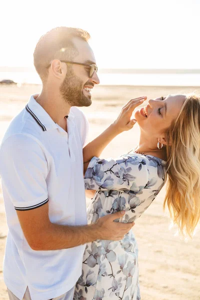 Foto de encantado casal bonito rindo e abraçando enquanto caminhava na praia ensolarada — Fotografia de Stock