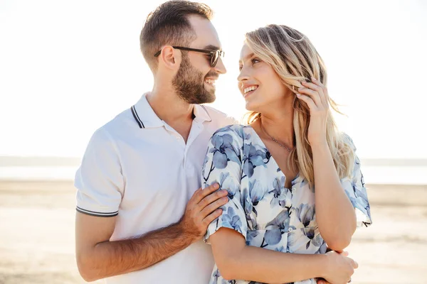 Фото красивої щасливої пари, яка посміхається один одному і обіймається під час прогулянки на сонячному пляжі — стокове фото