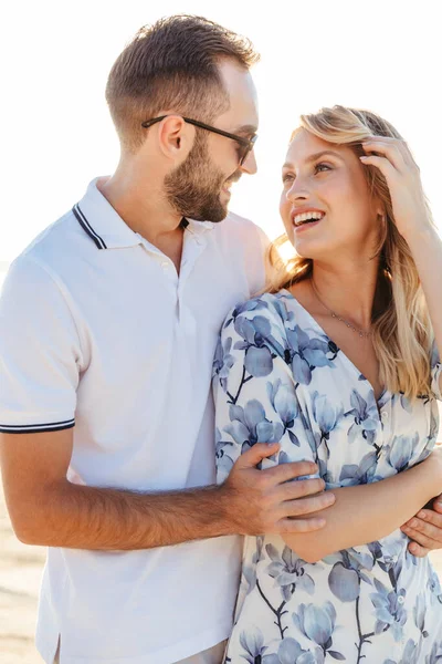 Kuva kauniista iloisesta pariskunnasta hymyilemässä toisilleen ja halaamassa kävellessään aurinkoisella rannalla — kuvapankkivalokuva