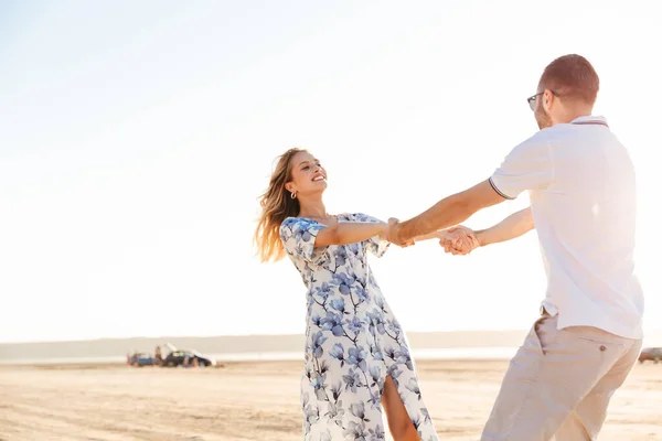 Foto de belo casal feliz sorrindo uns aos outros e rodopiando enquanto caminhava na praia ensolarada — Fotografia de Stock