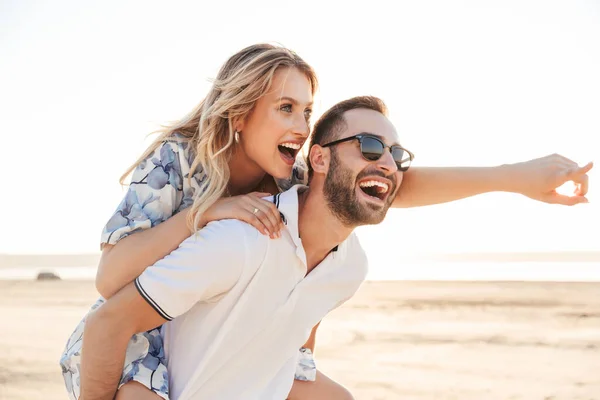 Foto de belo homem feliz sorrindo dando e cavalgando mulher sedutora enquanto caminhava na praia ensolarada — Fotografia de Stock