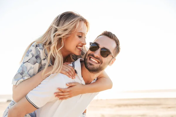 Foto do homem feliz sem barba sorrindo e dando passeio de piggyback mulher sedutora enquanto caminha na praia ensolarada — Fotografia de Stock