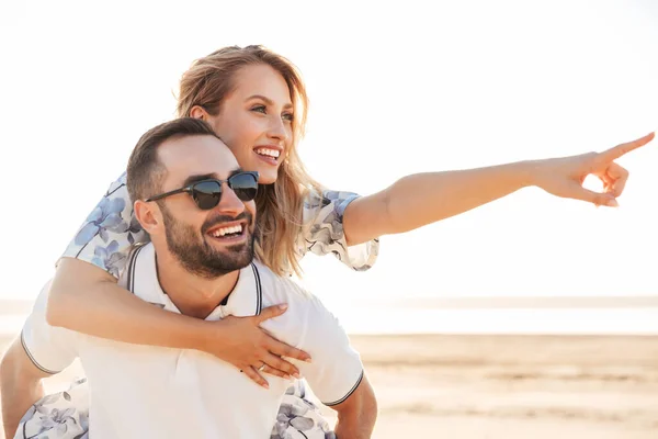 Foto van ongeschoren vrolijke man glimlachend en geven piggyback rijden verleidelijke vrouw tijdens het lopen op zonnig strand — Stockfoto