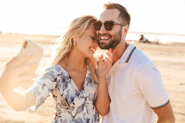 화창 한 해변을 걷고 있는 아름다운 커플이 웃으면서 핸드폰으로 셀카 사진을 찍는 모습 — 스톡 사진