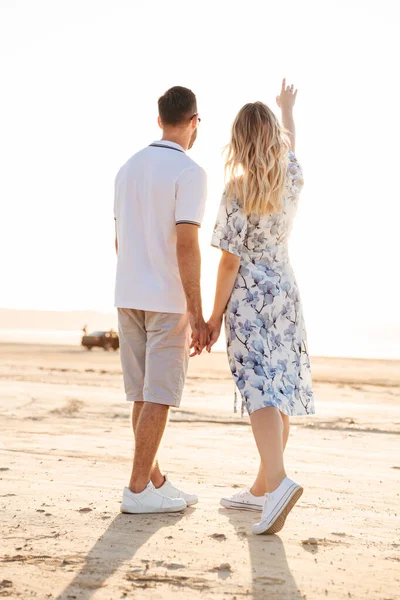 Foto de la parte posterior de la feliz pareja caucásica tomados de la mano mientras caminan en la playa soleada — Foto de Stock