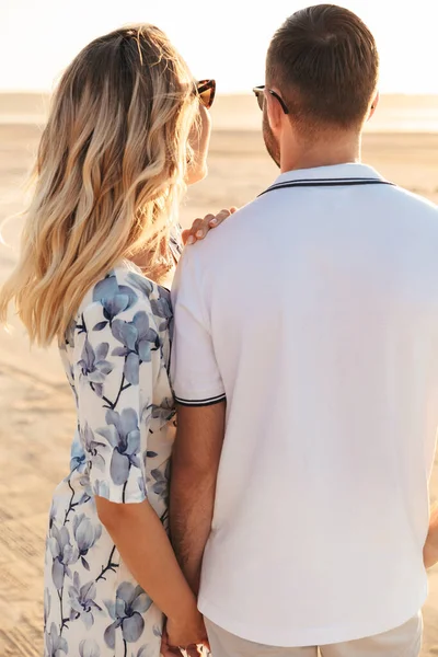 Photo de dos de caucasien beau couple tenant les mains ensemble tout en marchant sur la plage ensoleillée — Photo