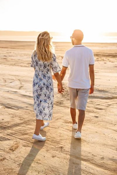 Güneşli sahilde yürürken el ele tutuşan beyaz tenli çekici çiftin fotoğrafı.