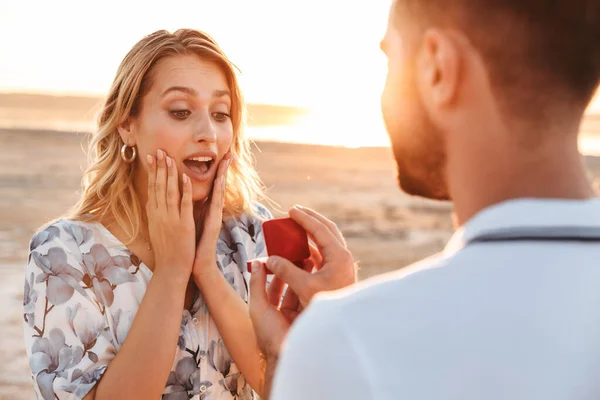 Foto de homem raspado fazendo proposta para sua mulher espantada com anel enquanto caminhava na praia ensolarada — Fotografia de Stock