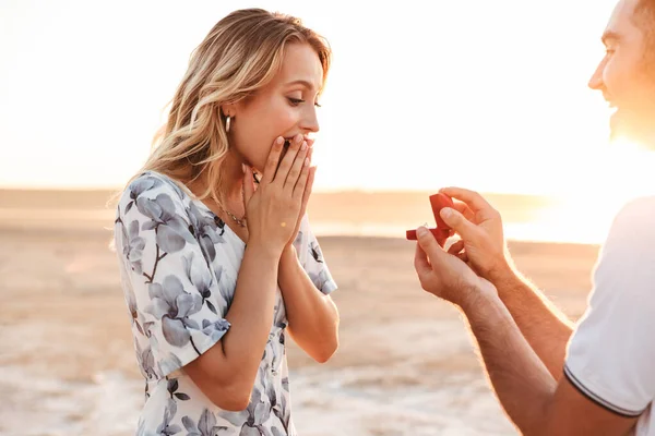 Foto av leende man som ger förslag till sin förbluffade kvinna med ring när du går på solig strand — Stockfoto