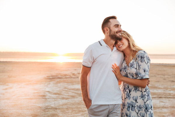 Фото счастливой молодой пары, улыбающейся и обнимающейся во время прогулки по солнечному пляжу
