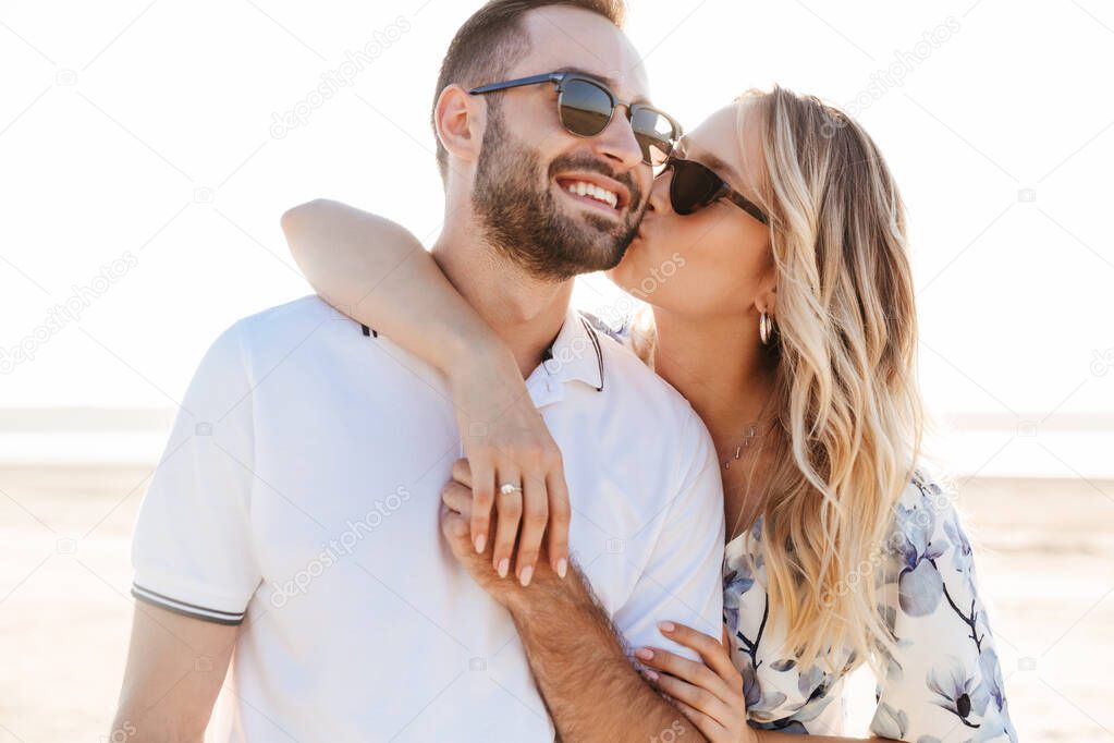 Photo of lovely young woman kissing and hugging handsome man while walking on sunny beach