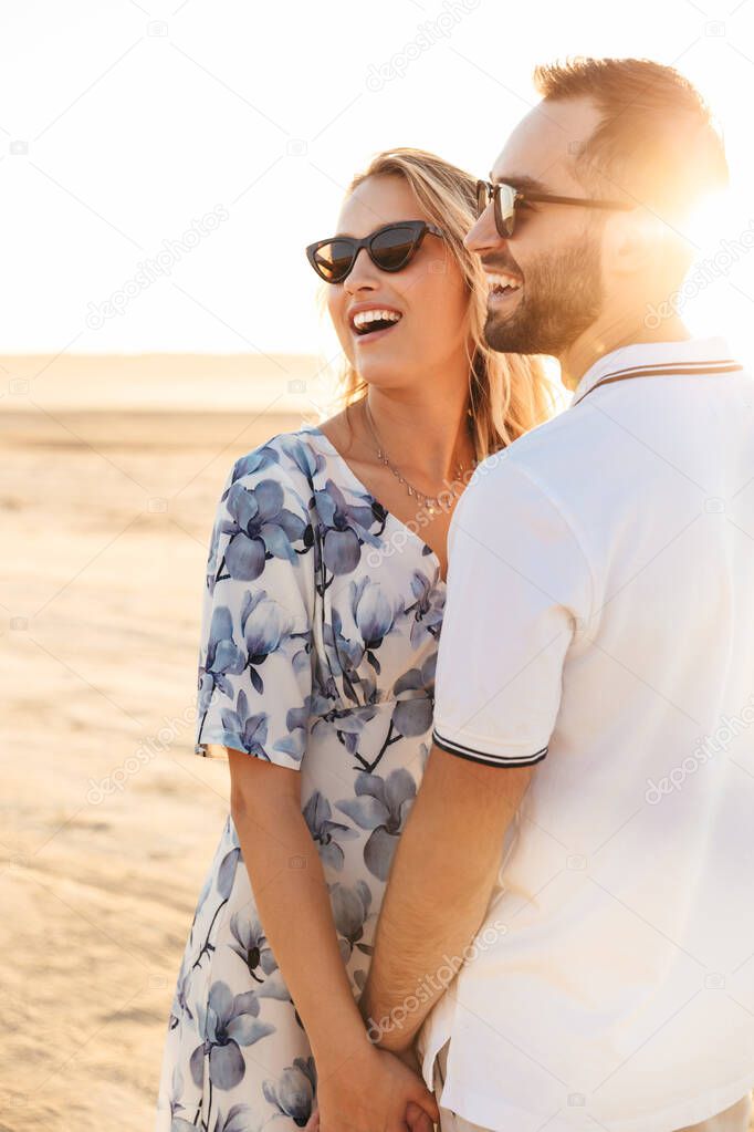 Photo of excited nice couple laughing and holding hands together while walking on sunny beach