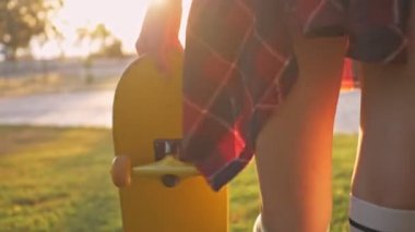 Parkta yürürken beyaz çoraplı genç kızın bacaklarını yakından izle. Parkta çimenlerde yürürken elinde kaykayla gülümseyen genç bir kızın yakın görüntüsü.