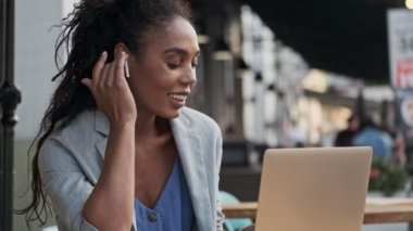 Mutlu Afrikalı genç iş kadını kulaklıklarla müzik dinliyor ve kafede otururken dizüstü bilgisayarla çalışıyor.