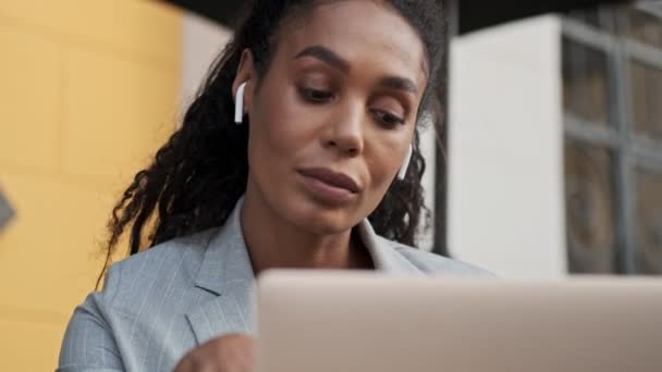 深刻な若いアフリカのビジネス女性のビューを閉じますイヤホンでラジオを聞いて カフェでラップトップで作業している間誰かを待っています — ストック動画