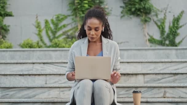 階段で屋外に座っている間 ラップトップで働いている耳ポッドを持つ若いアフリカのビジネス女性を満足させました — ストック動画