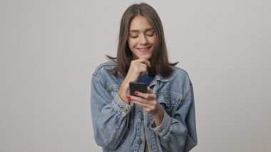 Gri arka plan üzerinde akıllı telefon kullanırken kot ceketli, gülümseyen ve çenesine dokunan oldukça düşünceli bir genç esmer kadın.