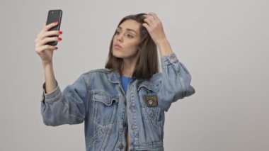 Kot ceketli, esmer, ciddi bir kadın. Gülümseyip saçını düzeltirken gri arka planda akıllı telefondan fotoğraf çekiyor.