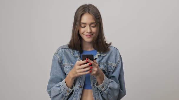 穿着牛仔夹克的快乐的年轻黑发女人一边笑一边用智能手机与灰色背景隔离 — 图库视频影像