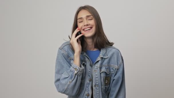穿着斜纹棉布夹克的快乐可爱的年轻黑发女人一边笑着 一边用智能手机与灰色背景隔绝地交谈 — 图库视频影像