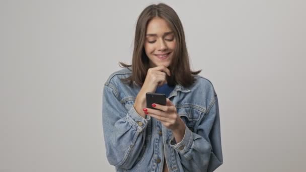 かなり思慮深い若いですブルネット女性でデニムジャケット笑顔とタッチ彼女のあごにスマートフォンを使用しています上のグレーの背景孤立 — ストック動画