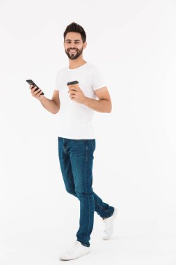 Kahvesini yudumlayan ve cep telefonuyla yazan mutlu bir adamın tam boy resmi.