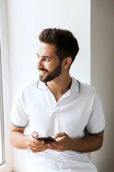 Pleased cheerful young happy man near window indoors using mobile phone. — Stock Photo, Image