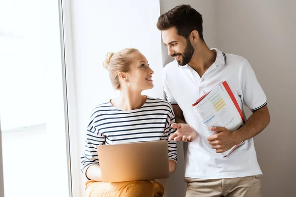 Young cheery happy loving couple friends man and woman near window indoors talking with each other holding copybooks using laptop computer. — Stock Photo, Image