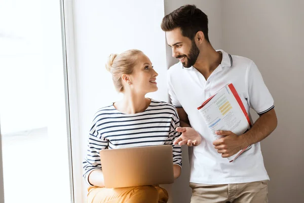 Joven alegre feliz pareja amorosa amigos hombre y mujer cerca de la ventana en el interior hablando entre sí celebración de libros de texto utilizando ordenador portátil . — Foto de Stock