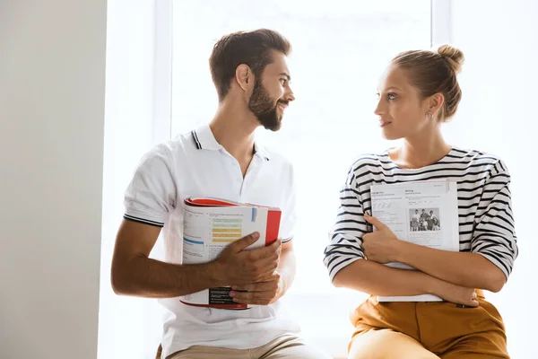 Joven pareja amorosa amigos hombre y mujer cerca de la ventana en el interior hablando entre sí celebración de copybooks . — Foto de Stock